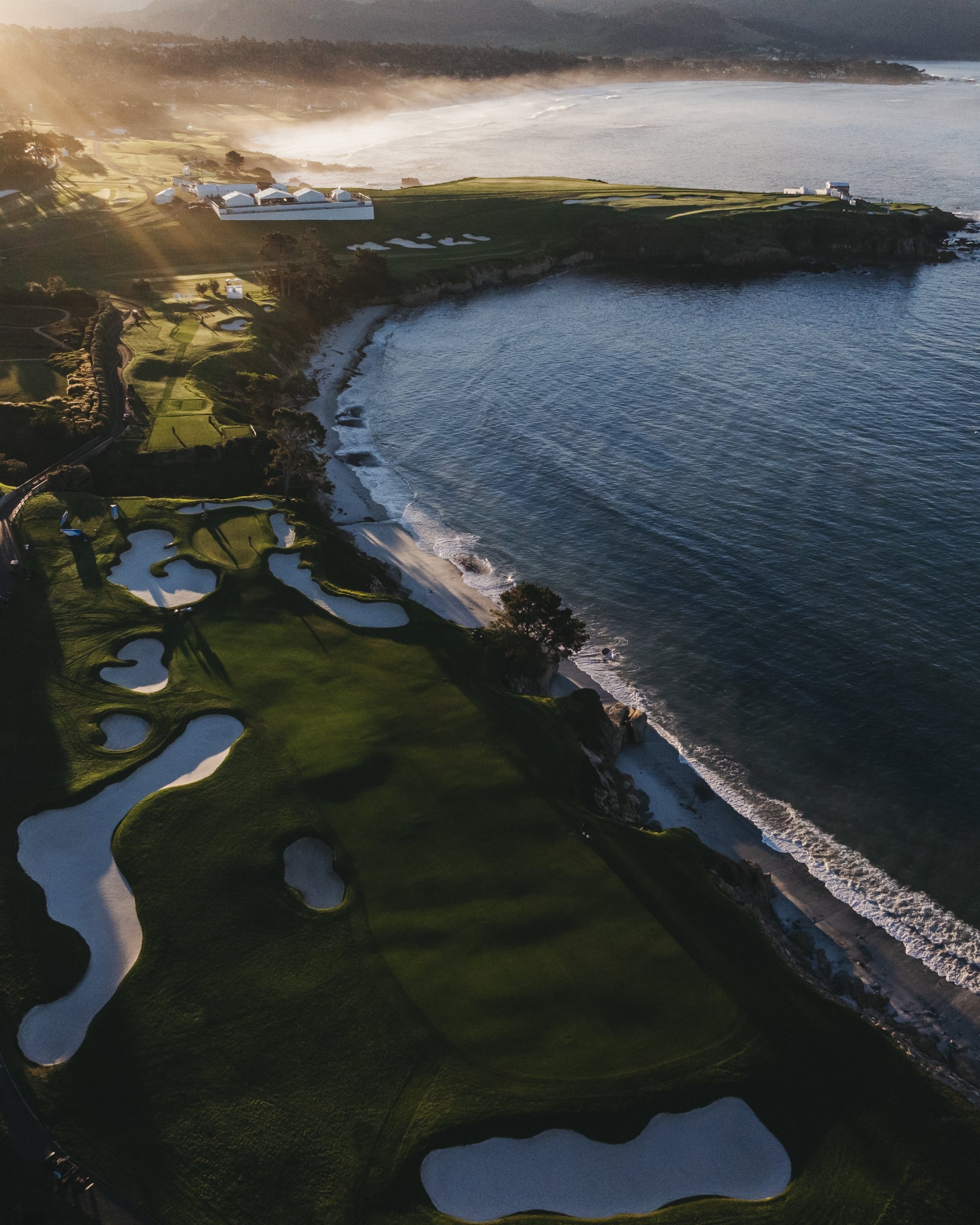 A dramatic, high-angle aerial view of the Pebble Beach golf course at sunrise (or sunset), with golden light rays beaming across the green fairways and cliffside sand bunkers, overlooking a sandy beach and the calm ocean.