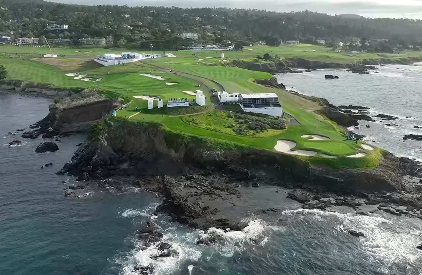 A wide aerial view of the Pebble Beach golf course on a dramatic rocky coastline, showing multiple holes, hospitality tents, and the ocean under a cloudy sky.