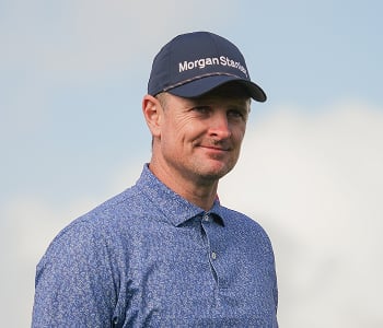 A headshot of golfer Justin Rose giving a slight smile, wearing a blue patterned polo shirt and a navy