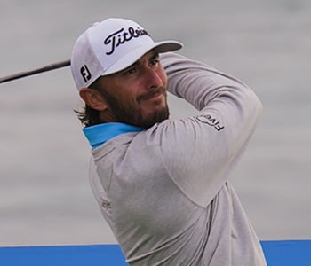 A close-up of golfer Max Homa in the middle of his swing, wearing a gray pullover with a blue collar and a white Titleist cap.