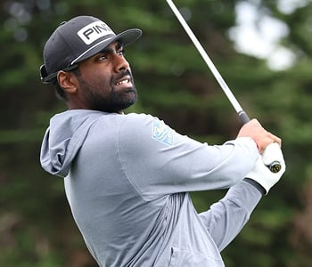 A close-up of golfer Sahith Theegala mid-swing, looking up intently, wearing a gray hooded sweatshirt and a black