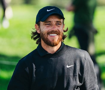 A close-up headshot of golfer Tommy Fleetwood smiling broadly, wearing a black Nike sweatshirt and a black Nike cap.
