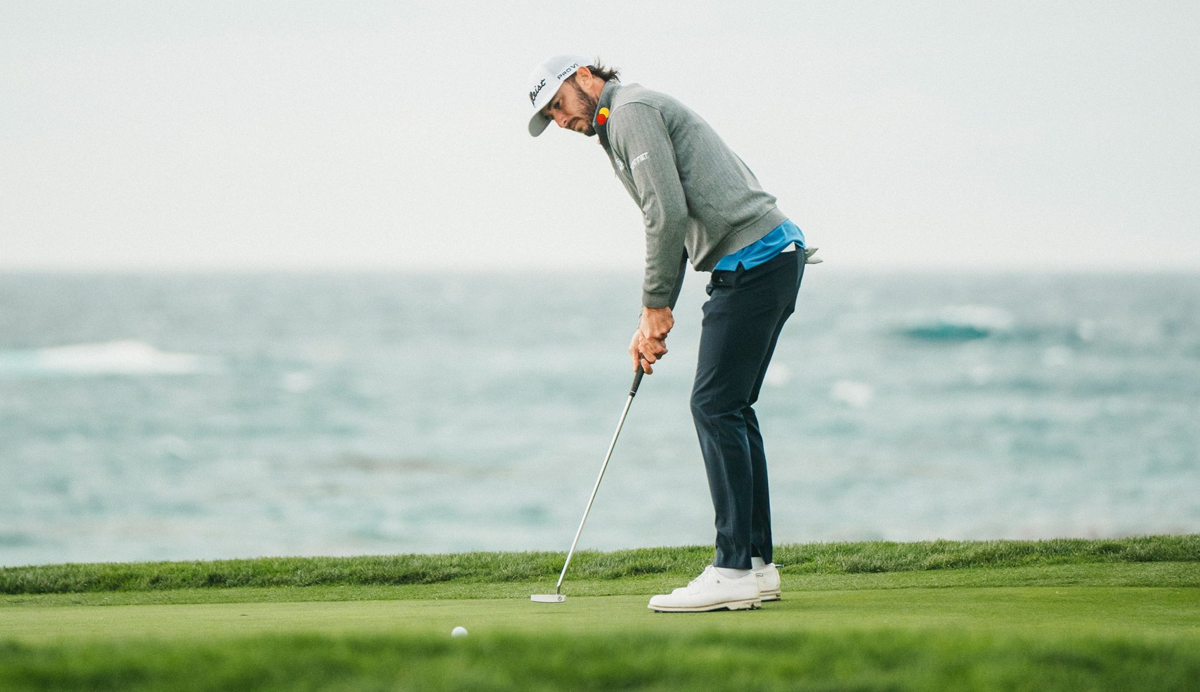 Golfer Max Homa (or similar player) bends over to line up a putt on a coastal green, with the rough ocean water serving as a dramatic background under a bright, cloudy sky.
