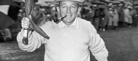 A black and white photo of Bing Crosby, wearing a golf cap and a light-colored sweater, smiling and holding an umbrella while biting on a pipe.