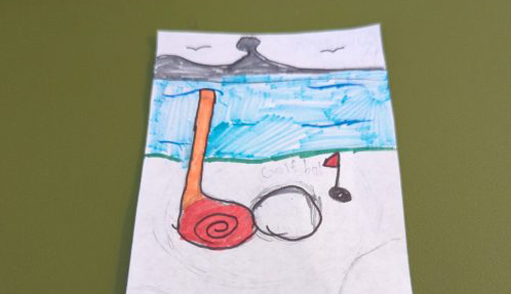 A child's drawing depicting a large orange golf club and a golf ball next to a flag on a sandy ground. The background is blue water and a dark mountain.