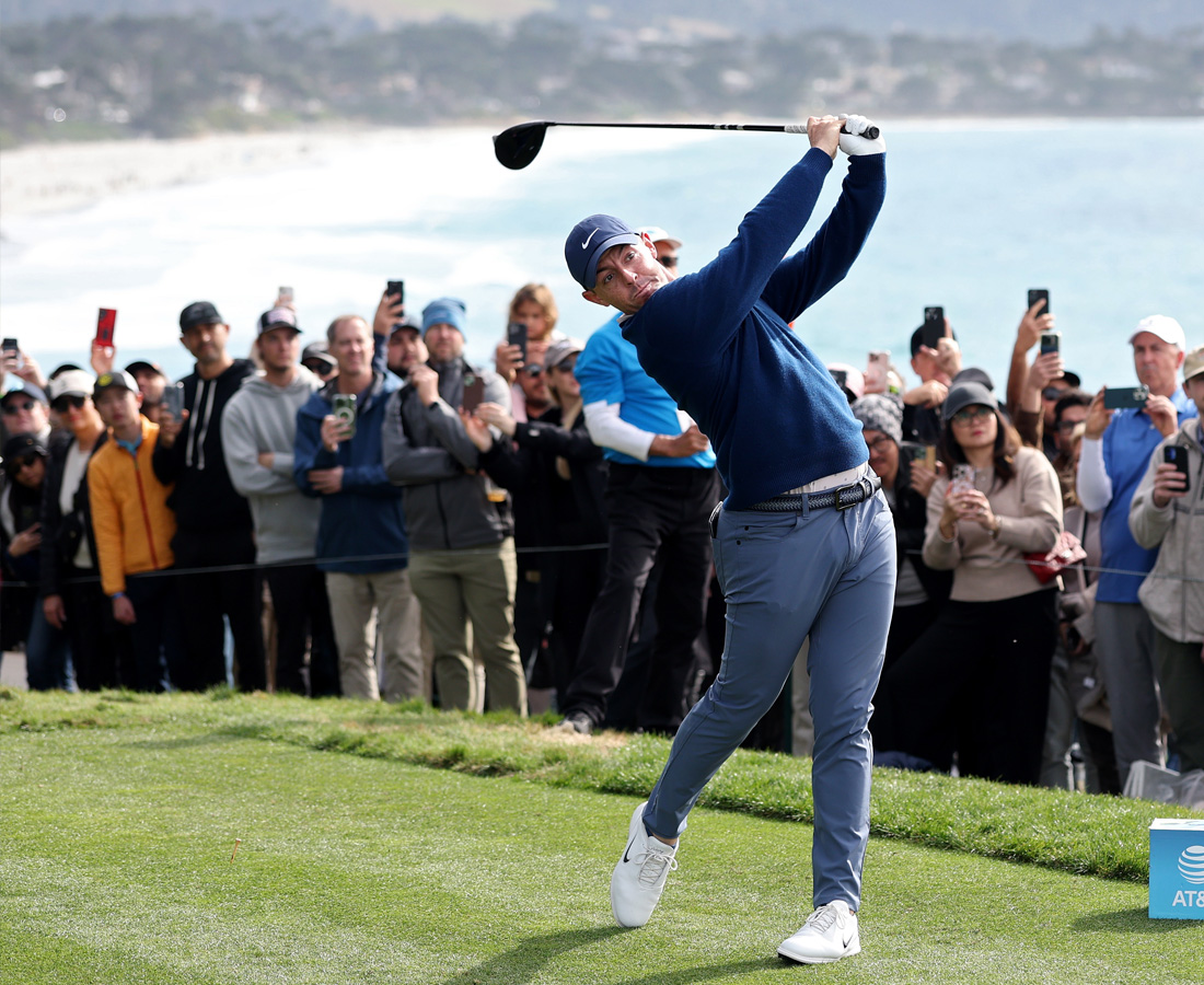 Golfer Rory McIlroy completes a powerful full-body swing from the tee box, with a large gallery of spectators behind him capturing the shot on their phones, and the ocean in the background.