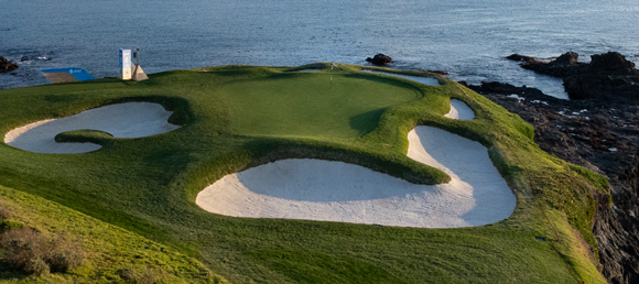 A dramatic view of a coastal golf green with four large, beautifully contoured sand bunkers. The green sits on a cliff overlooking the dark ocean water, with a small scoreboard structure visible.
