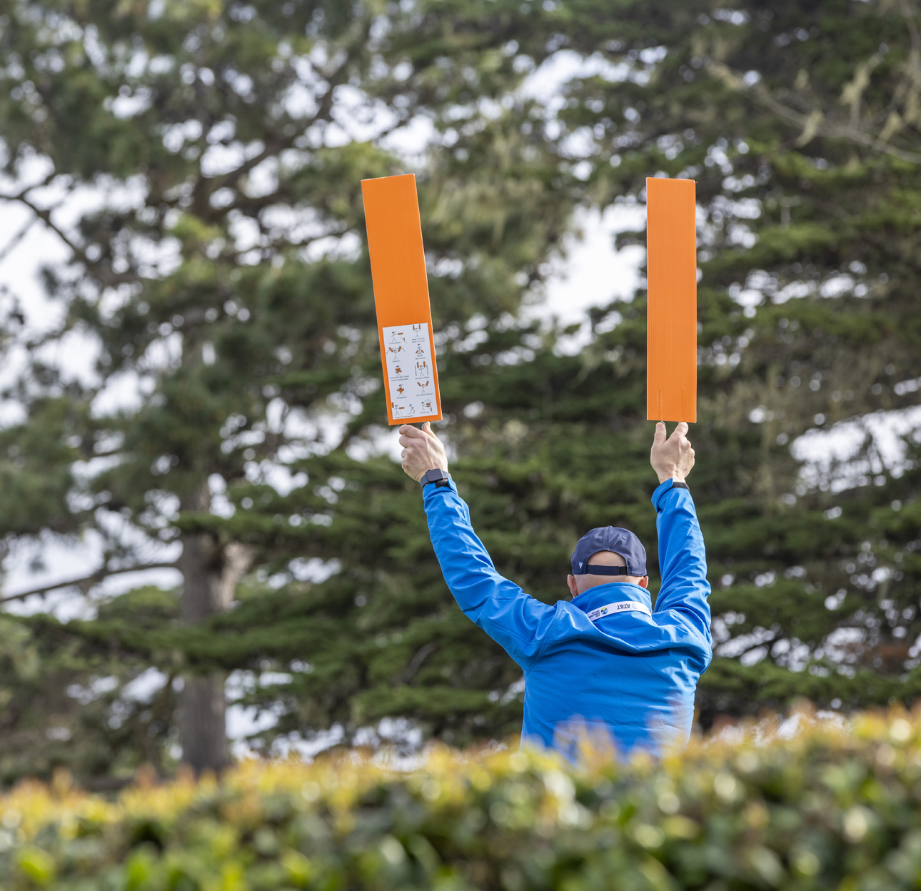 A golf course marshal (volunteer) in a blue jacket, seen from behind, holds two orange signal boards high above their head against a background of tall green trees.