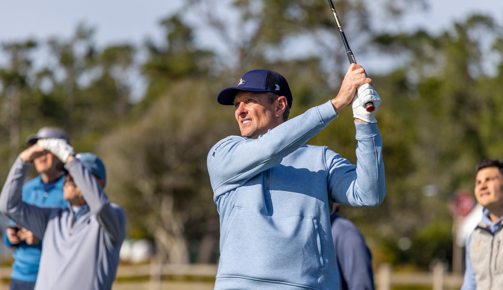Golfer Justin Rose (or similar player) is mid-swing on a sunny day, wearing a light blue pullover and a dark blue cap, with spectators watching behind him.