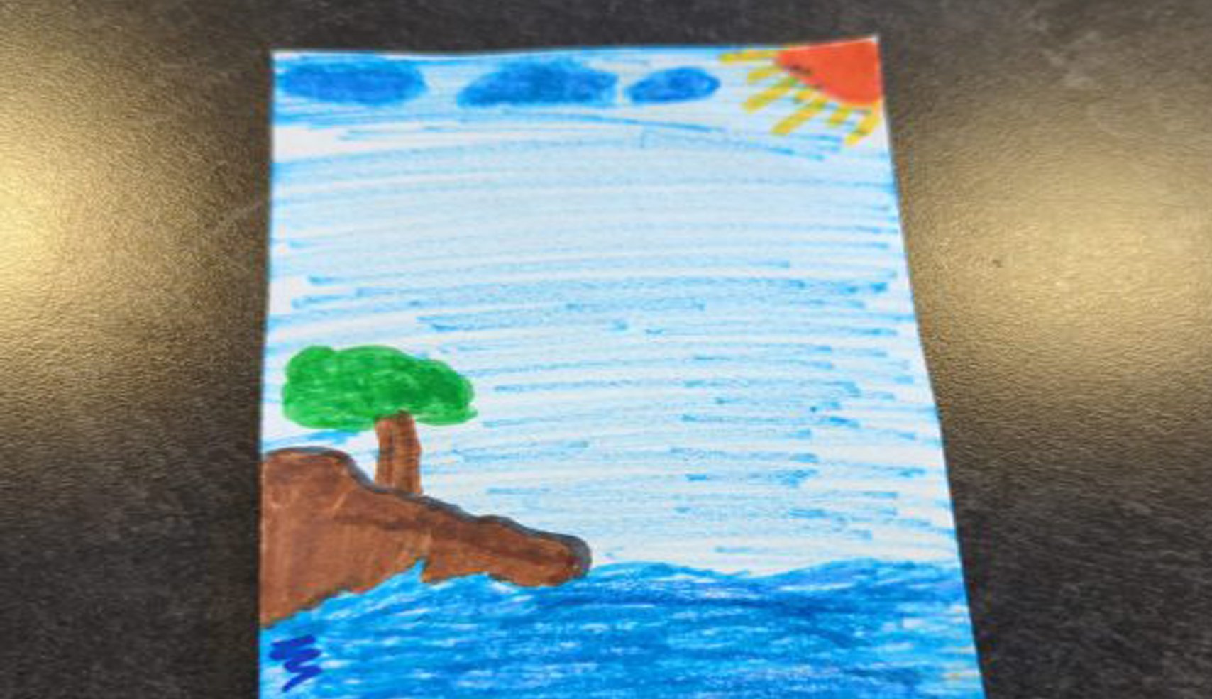 A child's drawing with a green-leafed tree growing on a brown cliff overlooking the blue ocean under a light blue sky with a red and yellow sun.