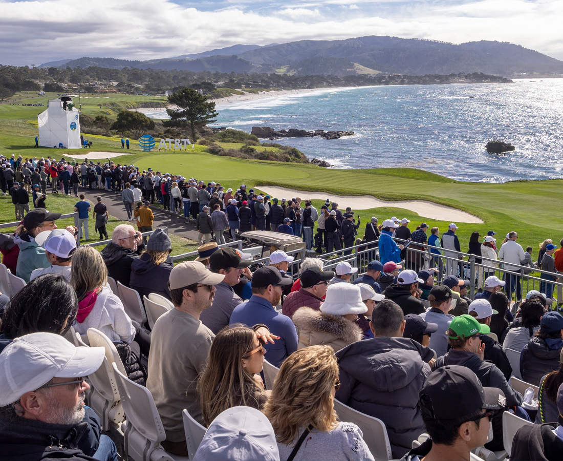 A wide view from the grandstands of the Pebble Beach course, showing a large crowd of spectators and the green fairway leading to the bright blue ocean, with mountains on the horizon.