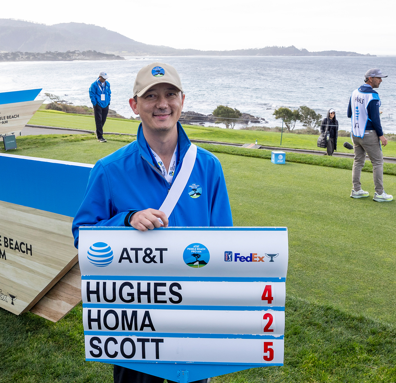 A smiling man in a blue volunteer jacket and beige cap holds a mobile scoreboard sign showing scores for Hughes, Homa, and Scott, with the ocean visible behind him.