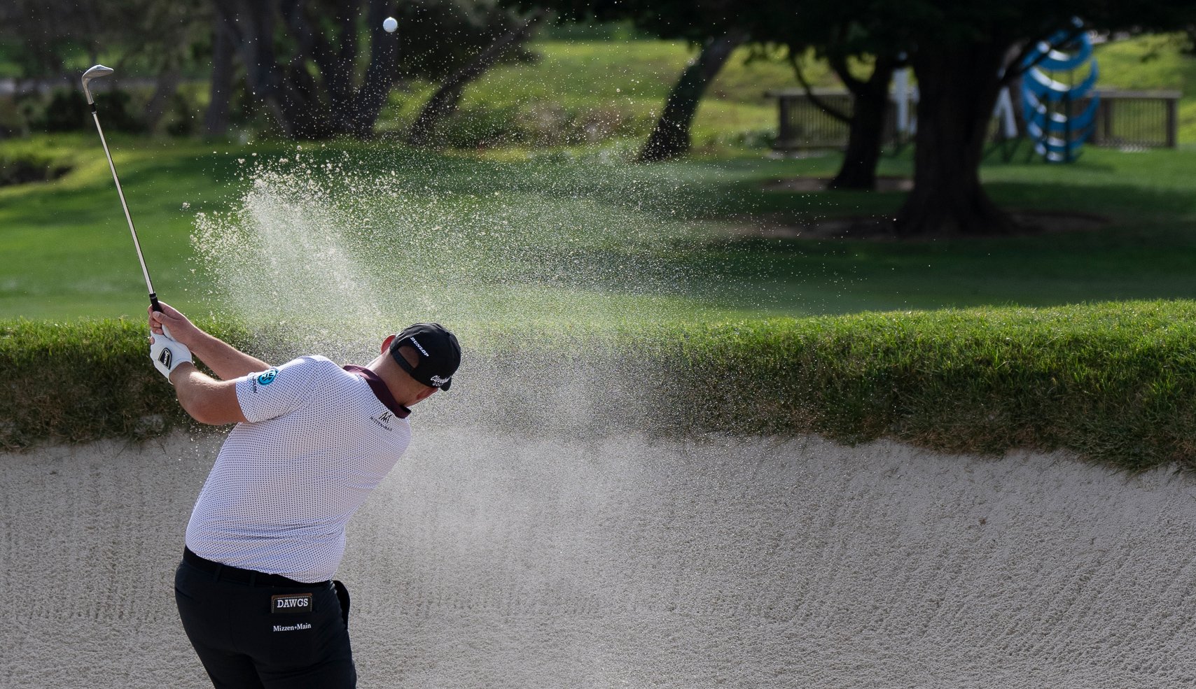 A golfer hits a shot out of a large sand bunker, kicking up a wide spray of sand and dust with the ball visible in the air.