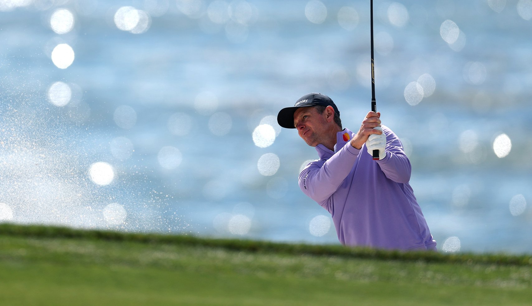A golfer in a lavender pullover hits a shot, with a large spray of sand to his left, framed against the brightly bokeh-lit ocean in the background.