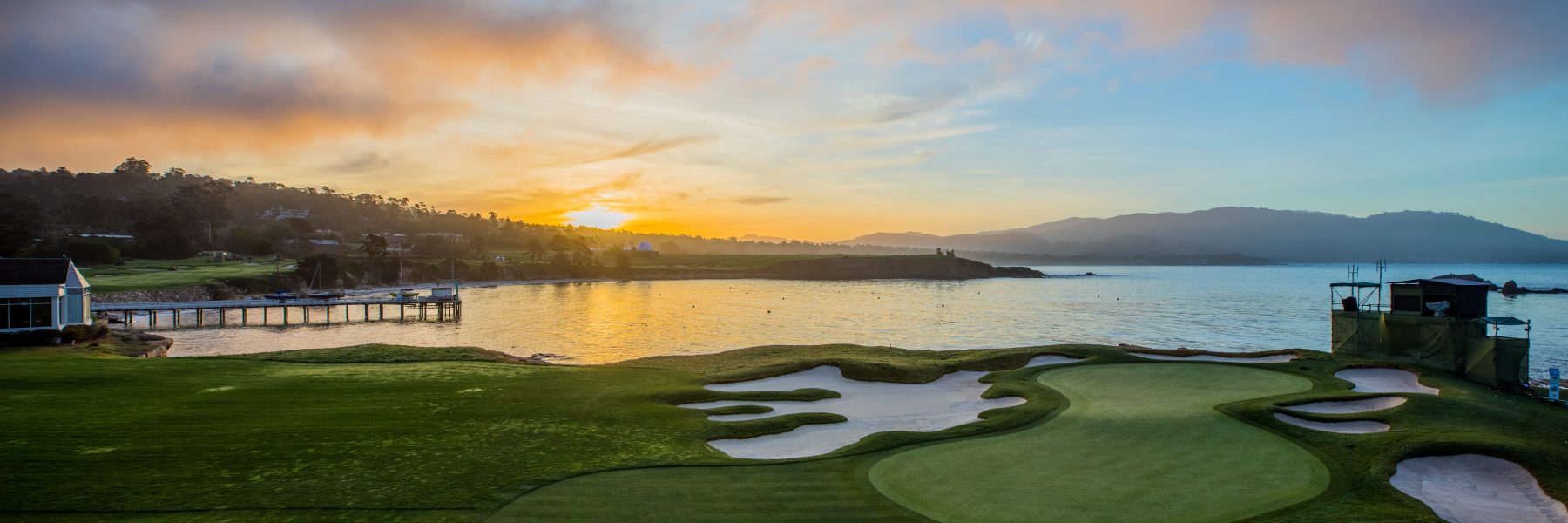A panoramic view of a golf green and sand traps at Pebble Beach at sunrise, with the ocean, a small pier, and distant hills visible in the golden light.