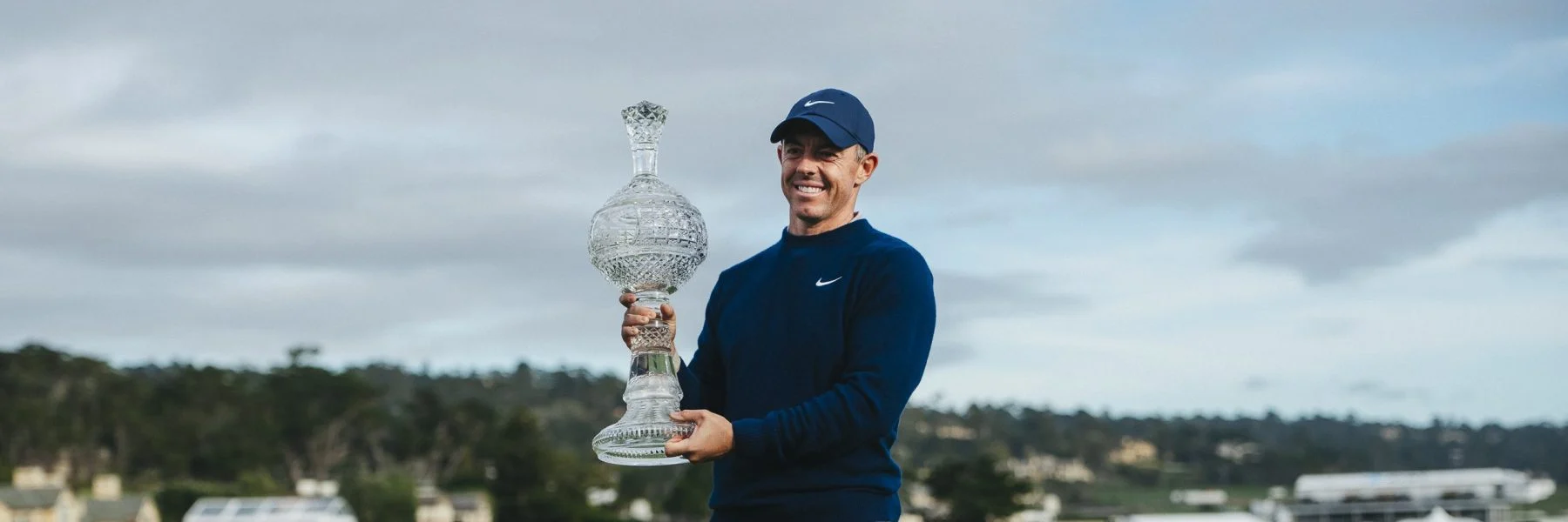 A wide horizontal shot of Rory McIlroy standing in the open, smiling and holding his large crystal trophy against a cloudy sky.