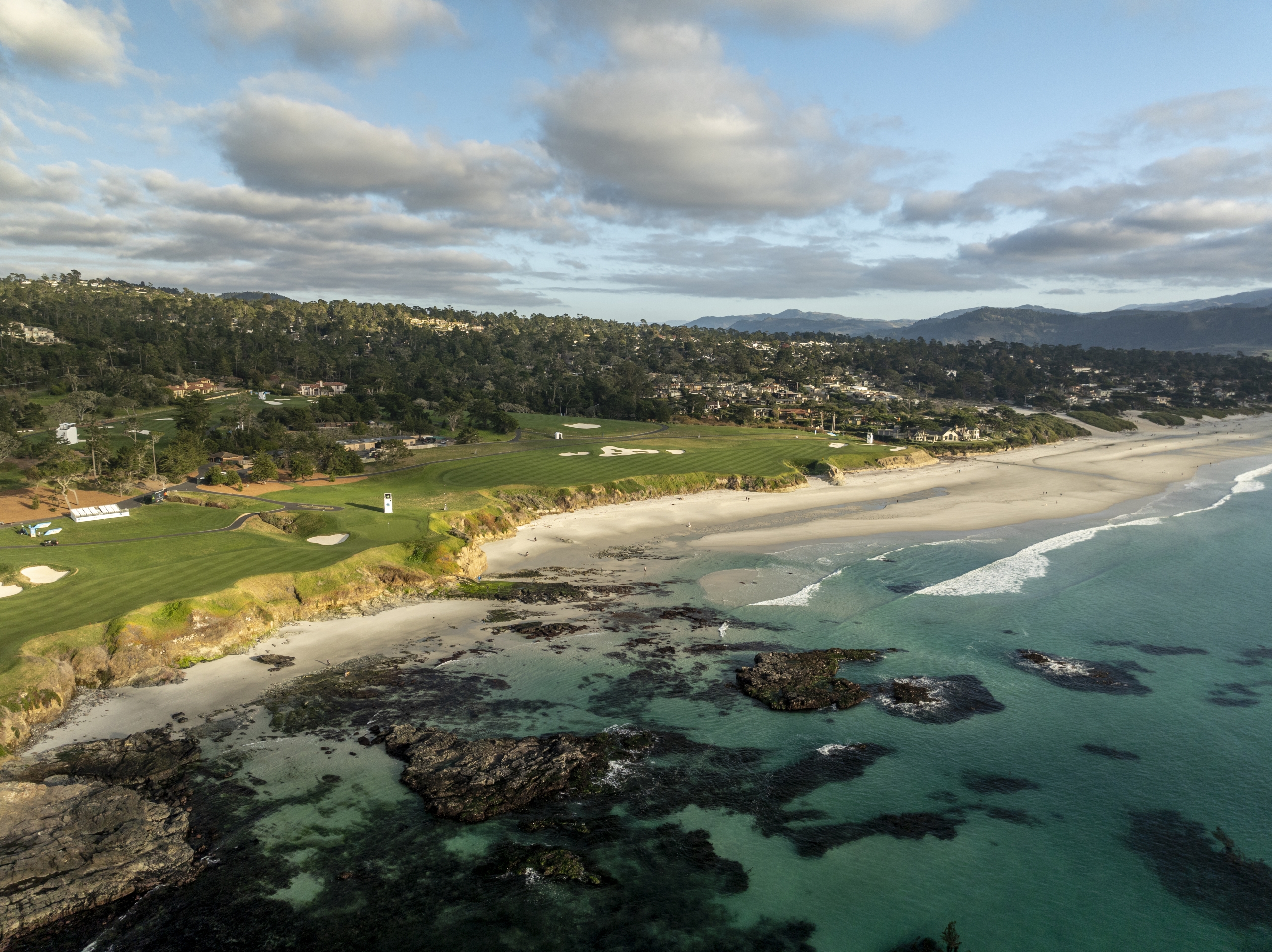 Aerial view of Pebble Beach's coastline, showing green fairways, a white sand beach, and turquoise ocean water.