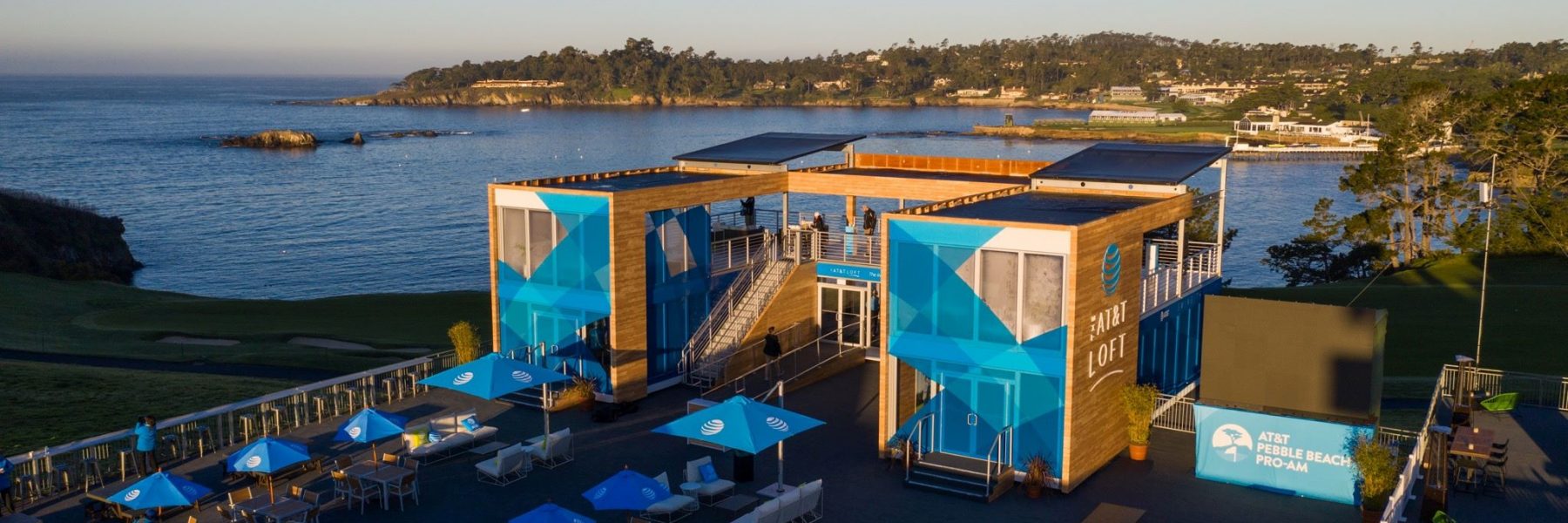 An aerial view of the modern, two-story AT&T Loft structure, constructed of wood and blue panels, overlooking the golf course and the ocean at sunset.