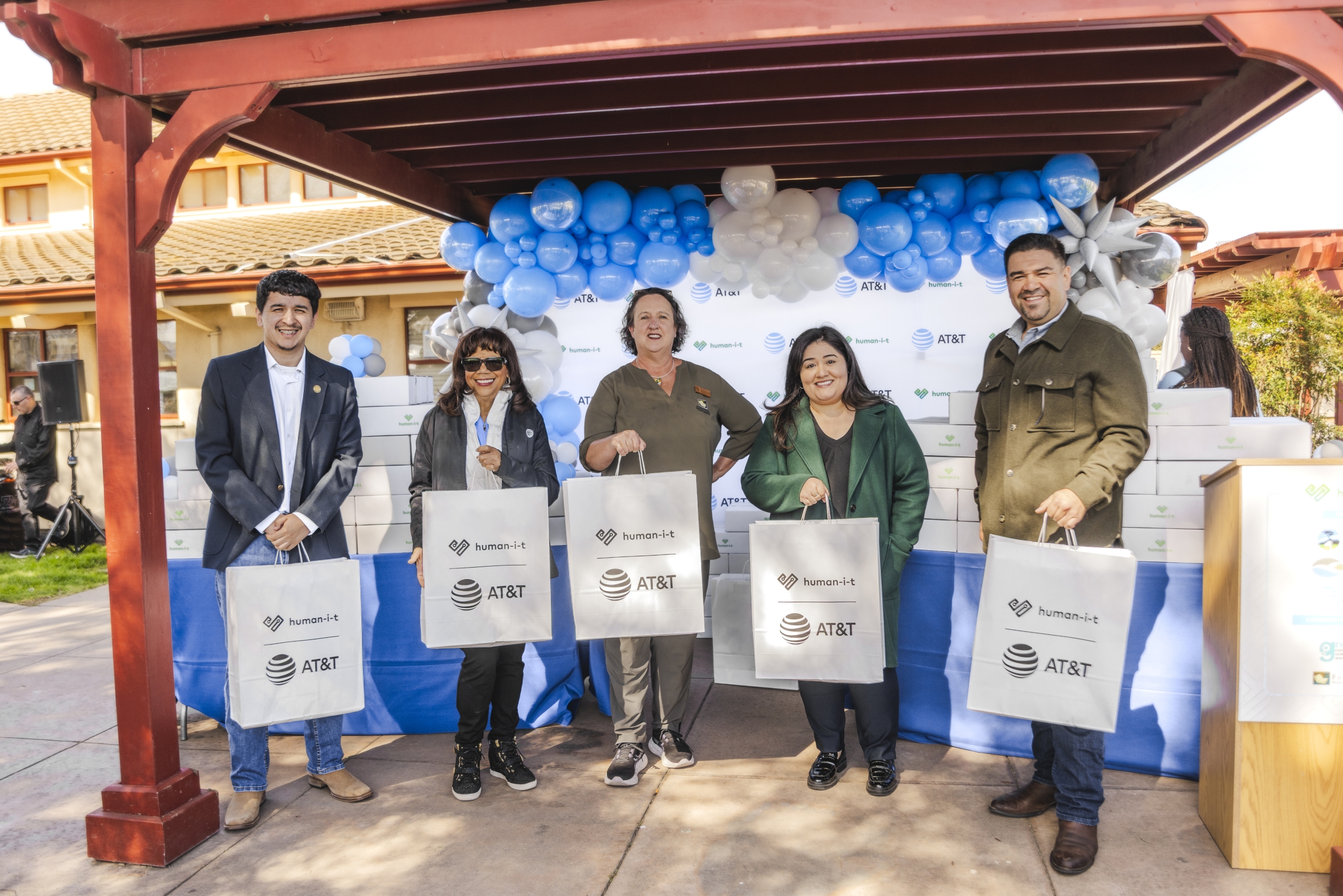 Five people, standing under an outdoor awning decorated with blue and white balloons, hold white shopping bags branded with AT&T and human-I-T (likely at a charitable event).