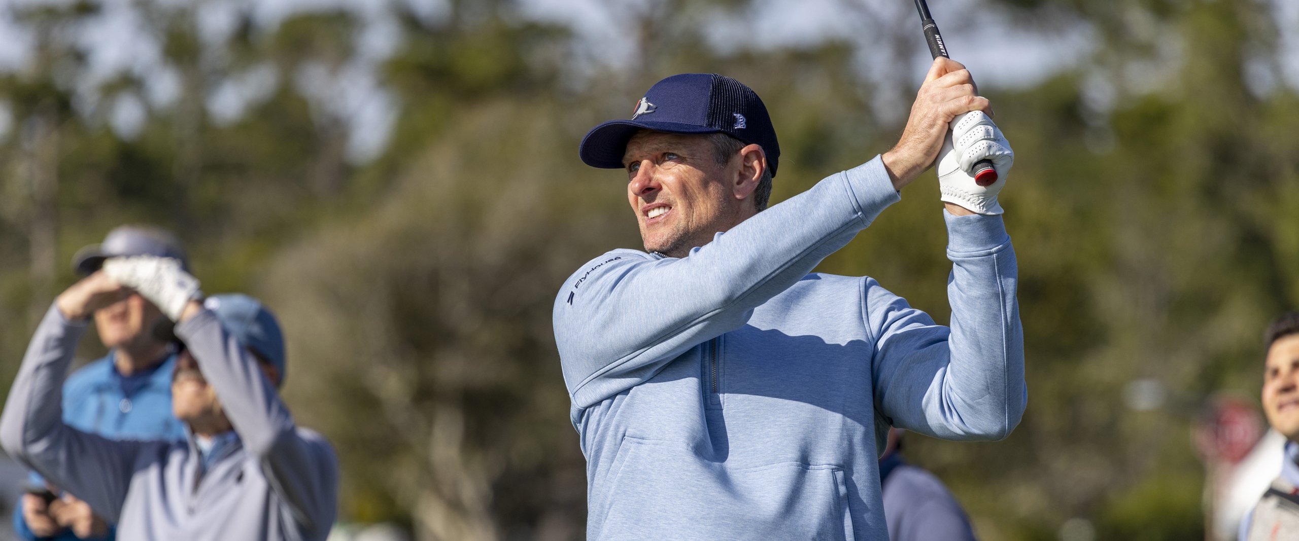 Golfer Justin Rose (or similar player) taking a swing, wearing a light blue pullover and a navy cap, with bright sunshine and blurred spectators in the background.