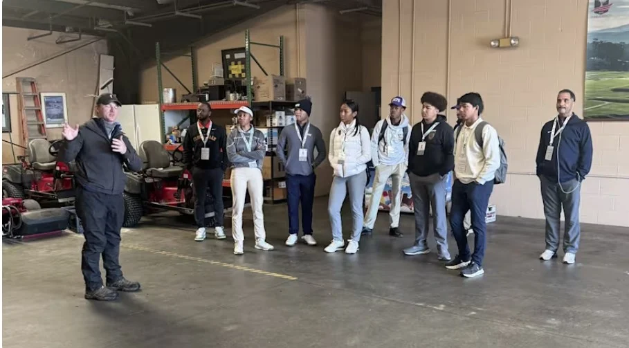 A man in a black jacket gestures as he speaks to a group of young students wearing event lanyards inside a large warehouse or maintenance garage.