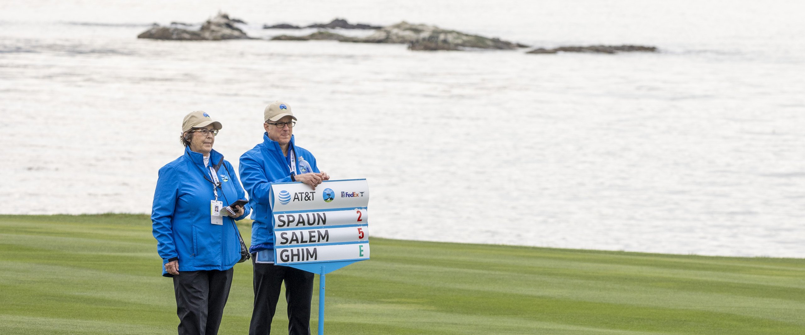 Two Pro-Am volunteers in blue jackets and beige caps stand next to a portable scoreboard on the green, with the ocean and rocks in the background under an overcast sky.