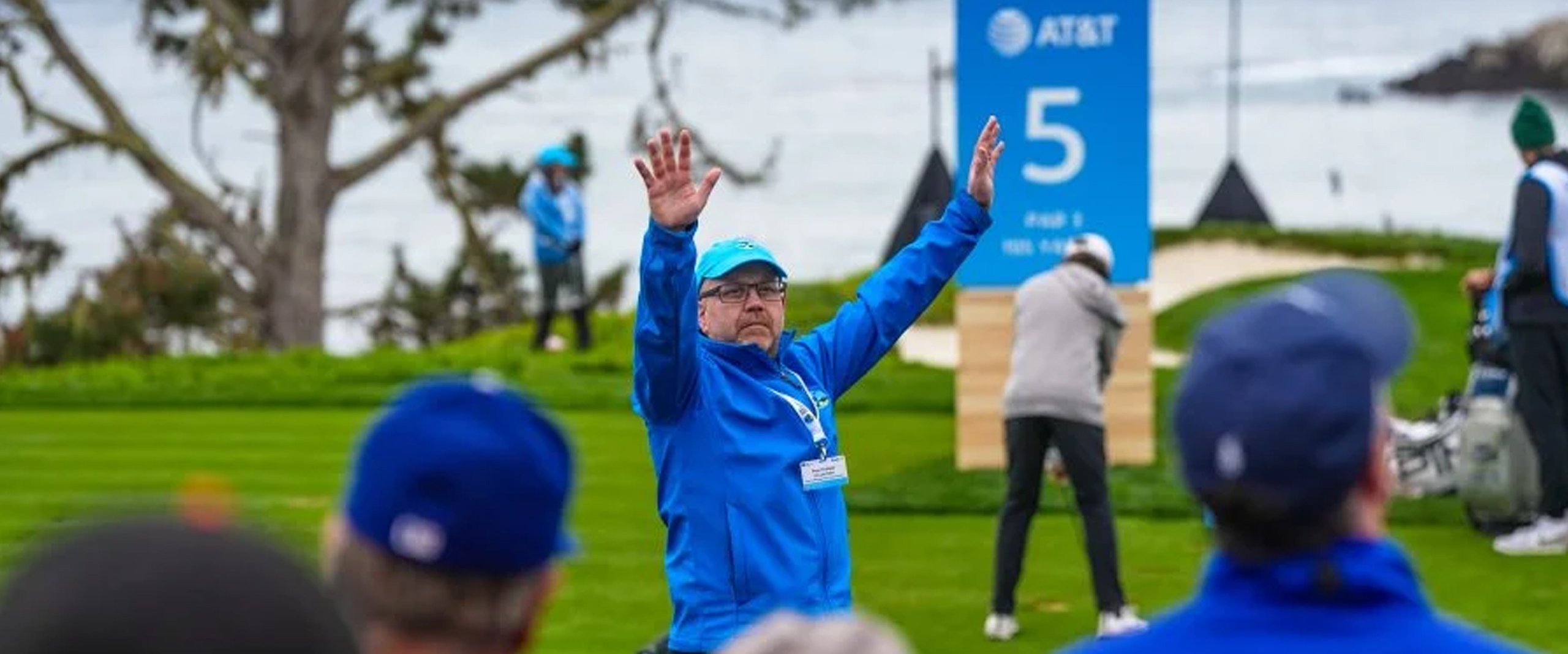 A close-up of an AT&T Pro-Am volunteer in a blue jacket with arms raised, signaling to the crowd. A large blue Hole 5 scoreboard is visible behind him.