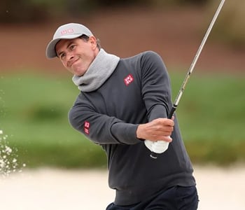 Professional golfer Adam Scott wearing a gray sweater and scarf, smiling as he hits a shot out of a sand bunker, kicking up a spray of sand.