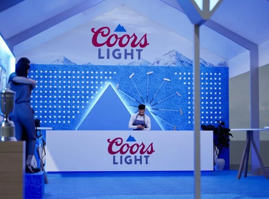 The interior of a Coors Light activation tent, featuring a bar counter with a bartender and a backdrop decorated with blue mountain imagery.