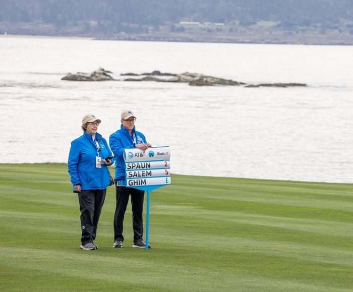 Two golf course volunteers in blue jackets and caps stand on the fairway, holding a portable scoreboard displaying the scores for Spaun, Salem, and Ghim, with the ocean in the background.