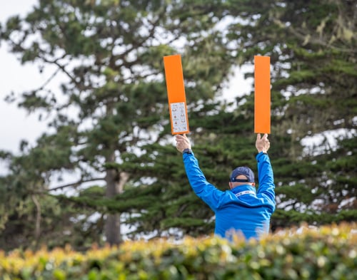 A golf course volunteer in a blue jacket, seen from behind, holding two bright orange signal boards up high against a background of pine trees.