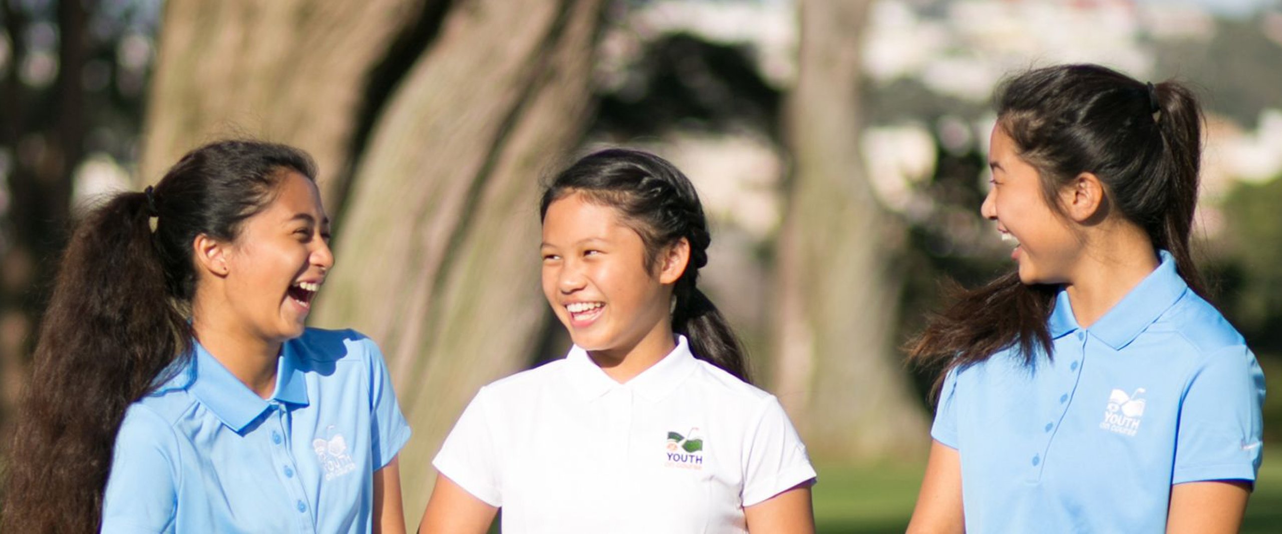 A close-up, horizontal image of three young girls (likely from a youth golf program) in matching polo shirts, smiling and laughing together with a natural background of trees.