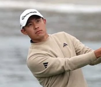 A headshot of professional golfer Collin Morikawa wearing a tan Adidas sweater and a white TaylorMade cap, looking intently toward the golf course.