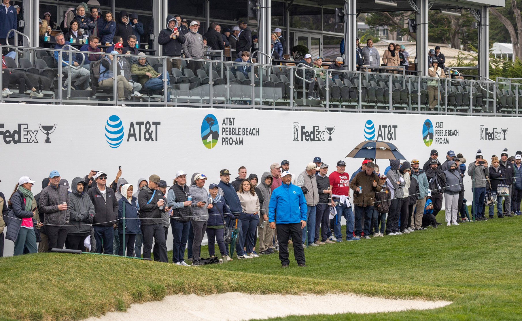 A crowd of spectators standing along the edge of the golf course near a sand bunker, watching the tournament action. Above them is a tiered seating structure with AT&T and FedEx branding.