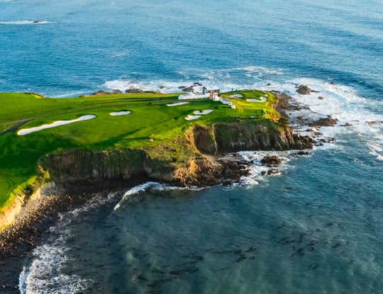 A high-angle aerial view of a coastal golf hole on a rocky cliff jutting into the ocean. The green is surrounded by white buildings and several sand bunkers, with crashing waves below.