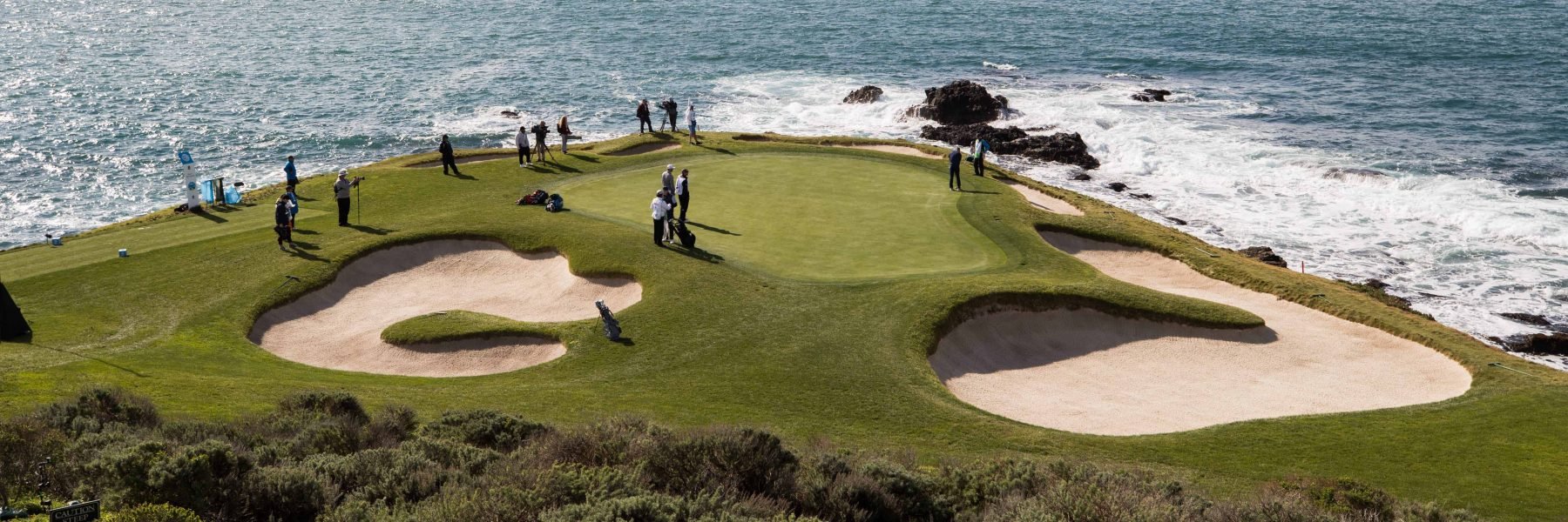 An iconic view of a golf green and two large sand bunkers on a cliffside overlooking the Pacific Ocean, with a group of people standing around the green.