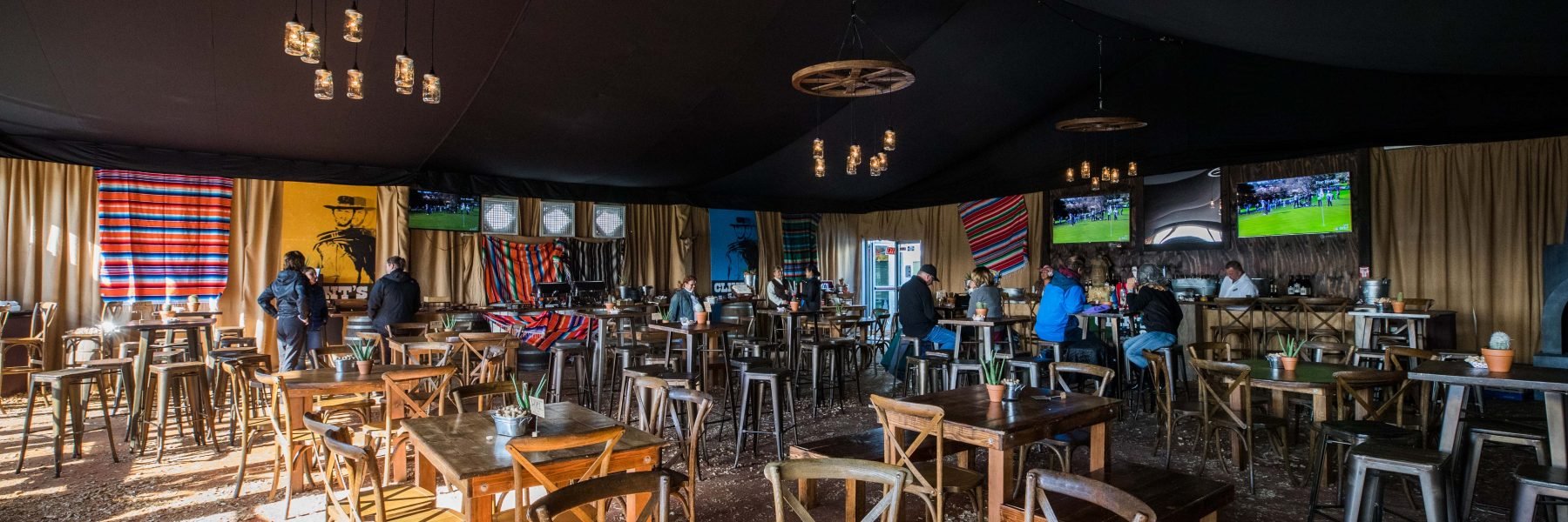 The interior of a rustic-themed hospitality tent or bar, featuring wooden tables and high chairs, with Mexican serapes (blankets) and TVs on the walls, giving a Western or Southwestern ambiance.