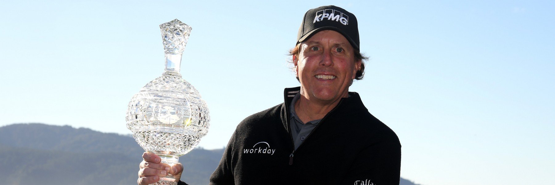 Professional golfer Phil Mickelson smiling and holding a large, ornate crystal trophy, wearing a black pullover and a KPMG hat, with a clear blue sky and distant hills behind him.
