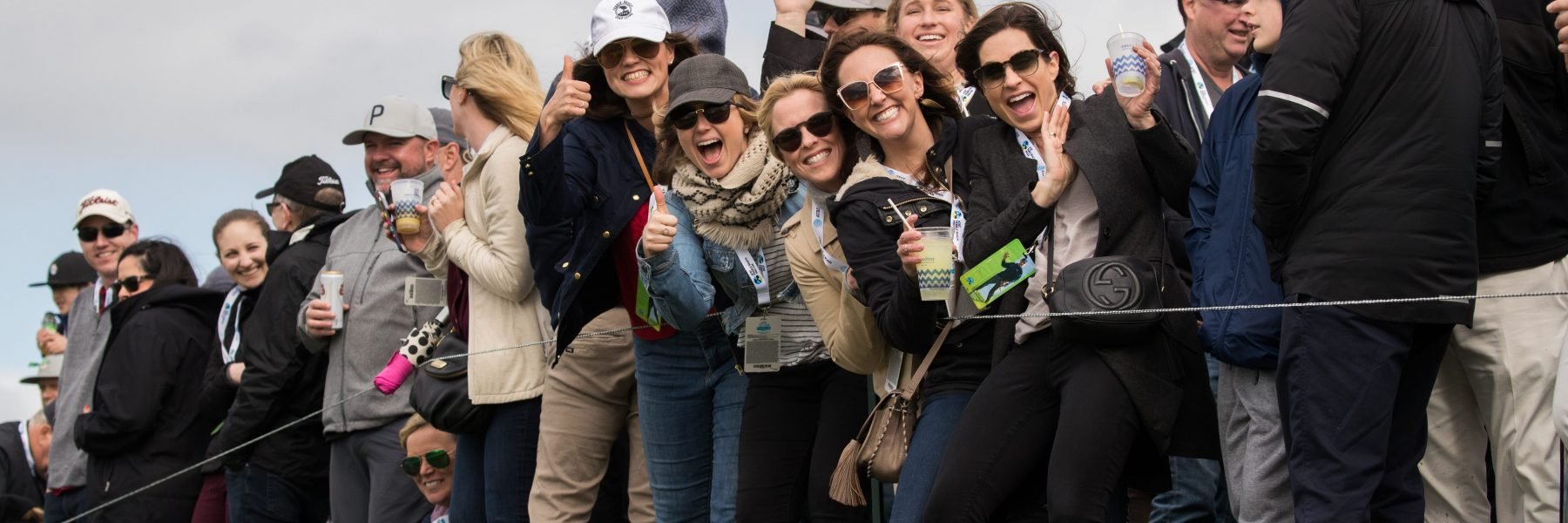 A wide, horizontal shot of a crowd of excited spectators smiling, cheering, and giving thumbs-up gestures as they watch the action on the golf course.