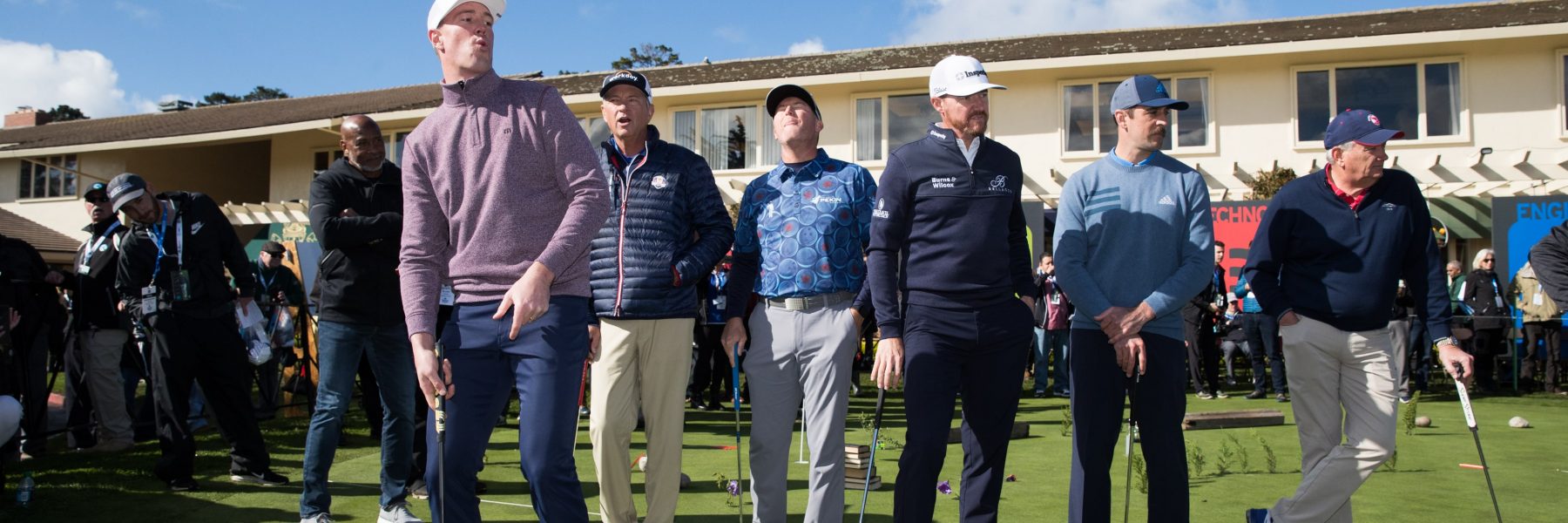 A group of several men, including professional golfers and amateurs, standing side-by-side on a putting green in front of a white building, watching a shot.