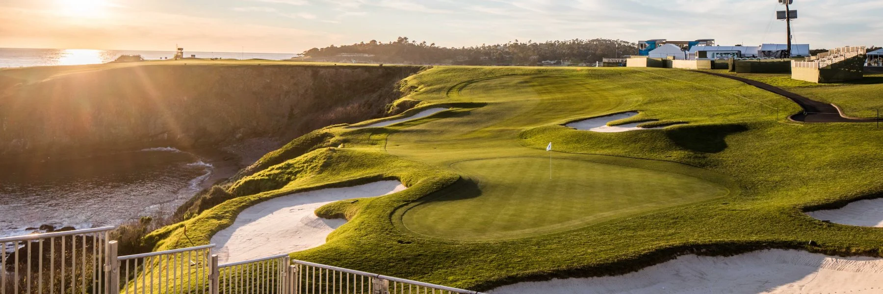A golf green and sand bunker on a cliff edge at Pebble Beach at sunset, with the golden light flaring over the course and the vast ocean and coastline in the background.