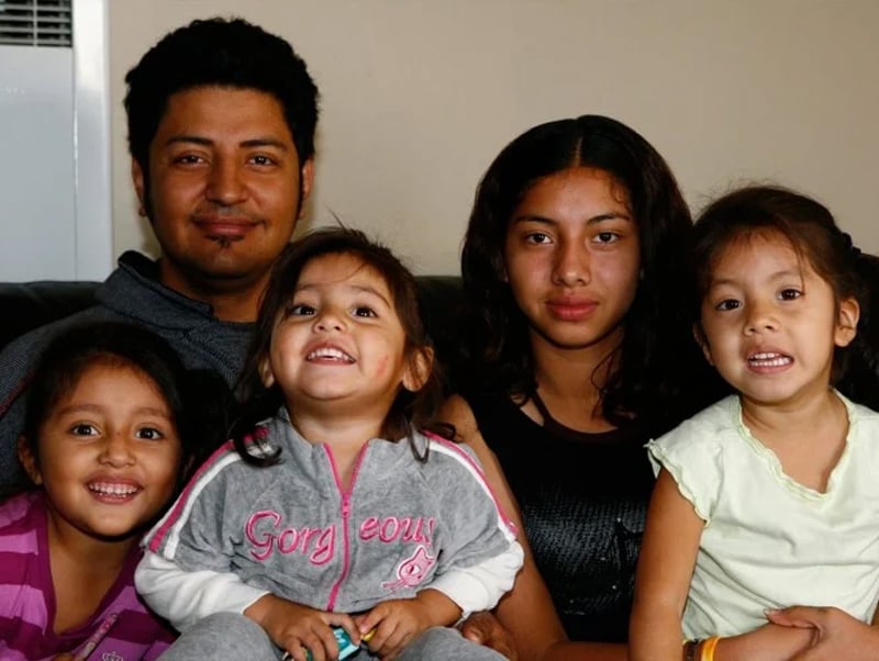 A close-up portrait of a Hispanic family, showing a father and four young girls sitting closely together, smiling at the camera.