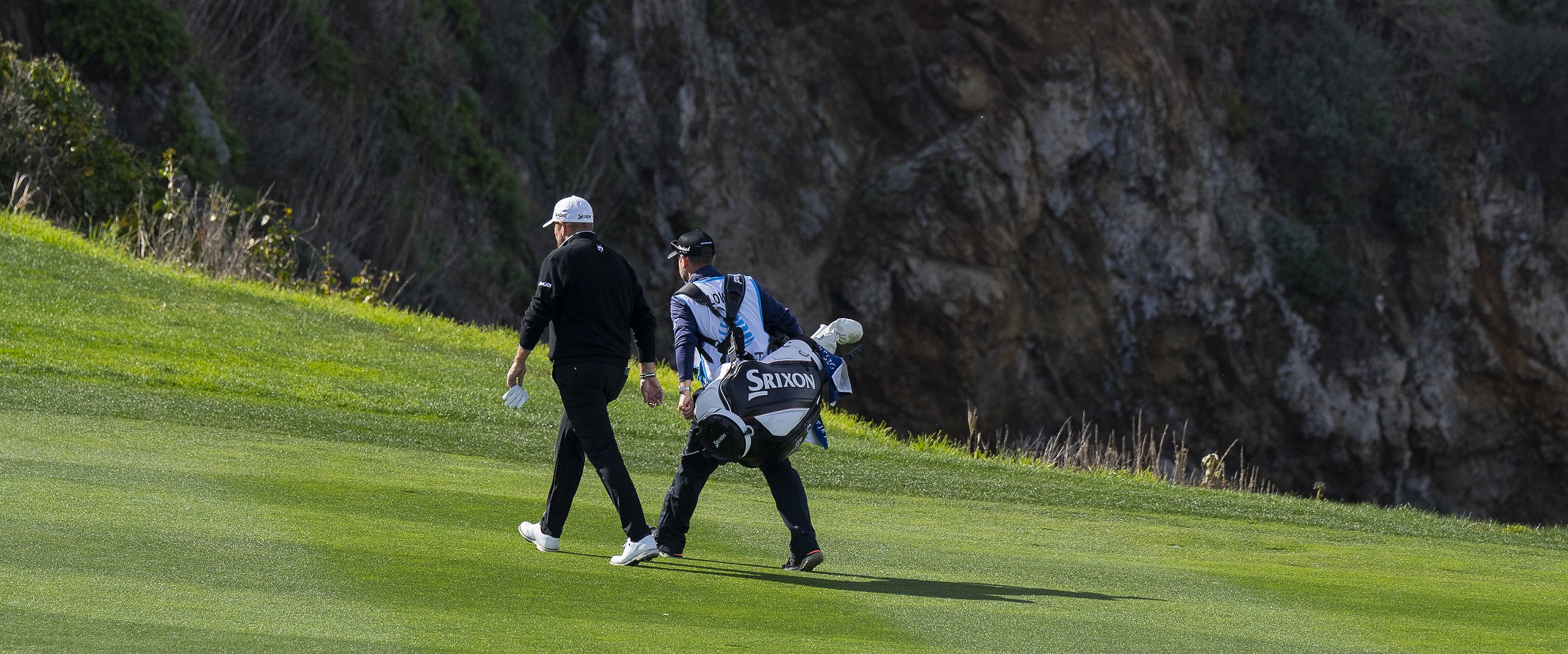 A golfer and his caddie (wearing a Srixon bag) walking away from the camera up a grassy hill towards a sheer cliff face.