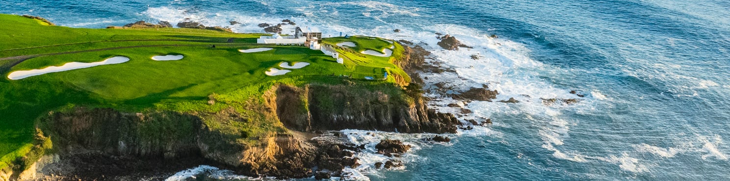 A wide aerial view of a dramatic coastal golf hole on a cliff, where green fairways meet a rocky shoreline and the deep blue ocean with white, foamy waves.