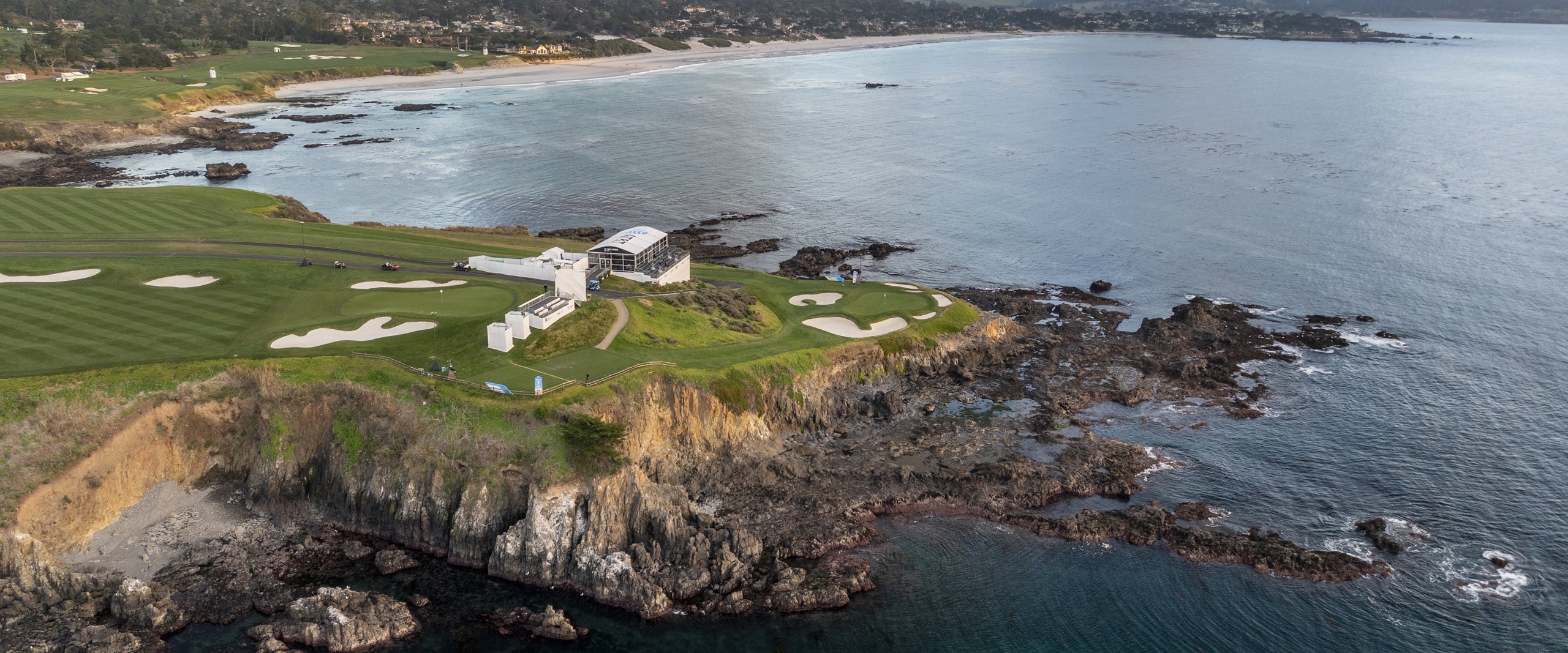 A breathtaking aerial view of the Pebble Beach coastline, showing the dramatic cliffs, crashing waves, and the golf course clinging to the edge, featuring a white hospitality structure overlooking the ocean.
