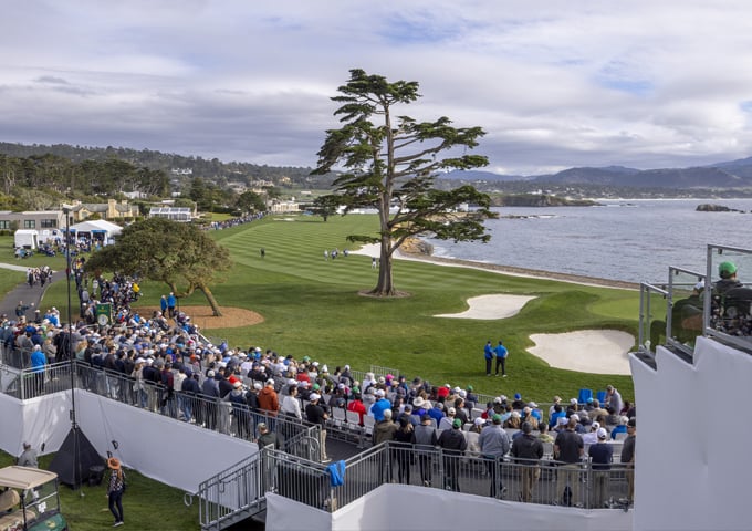 A wide view of the Pebble Beach course and coastline, showing spectators watching from a hillside and from elevated hospitality structures that sit directly above a dramatic, dark cliff face.