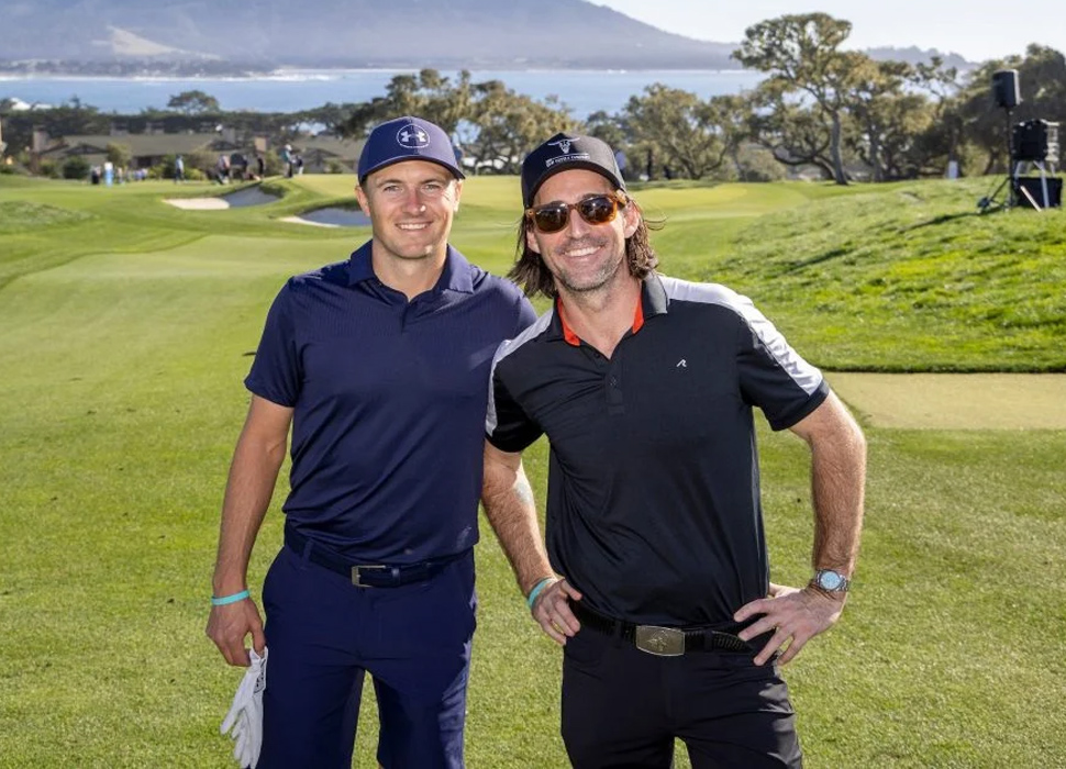 Professional golfer Jordan Spieth and an amateur partner (Jake Owen) pose side-by-side on the golf course, smiling, with the ocean and hills in the background.