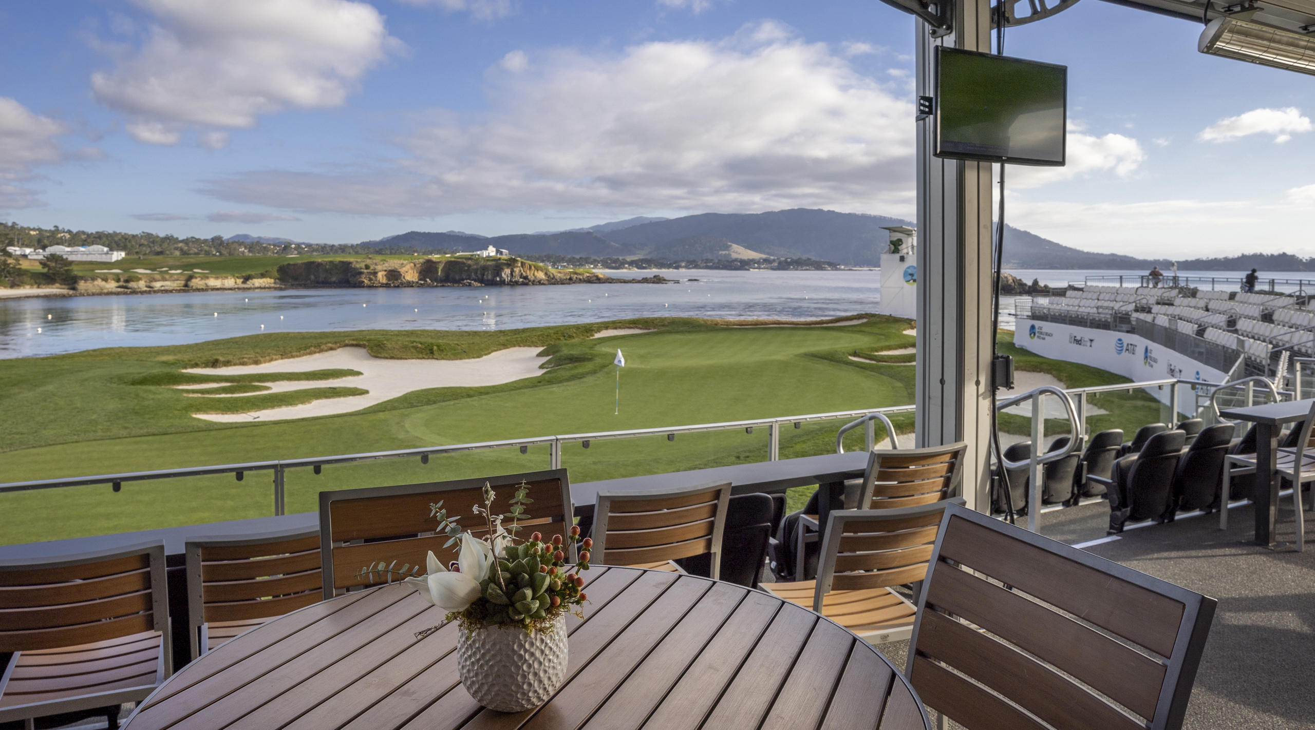 A view from a covered hospitality lounge featuring a wooden table setting and chairs. The window offers a beautiful view of a coastal golf green, the bay, and mountains.