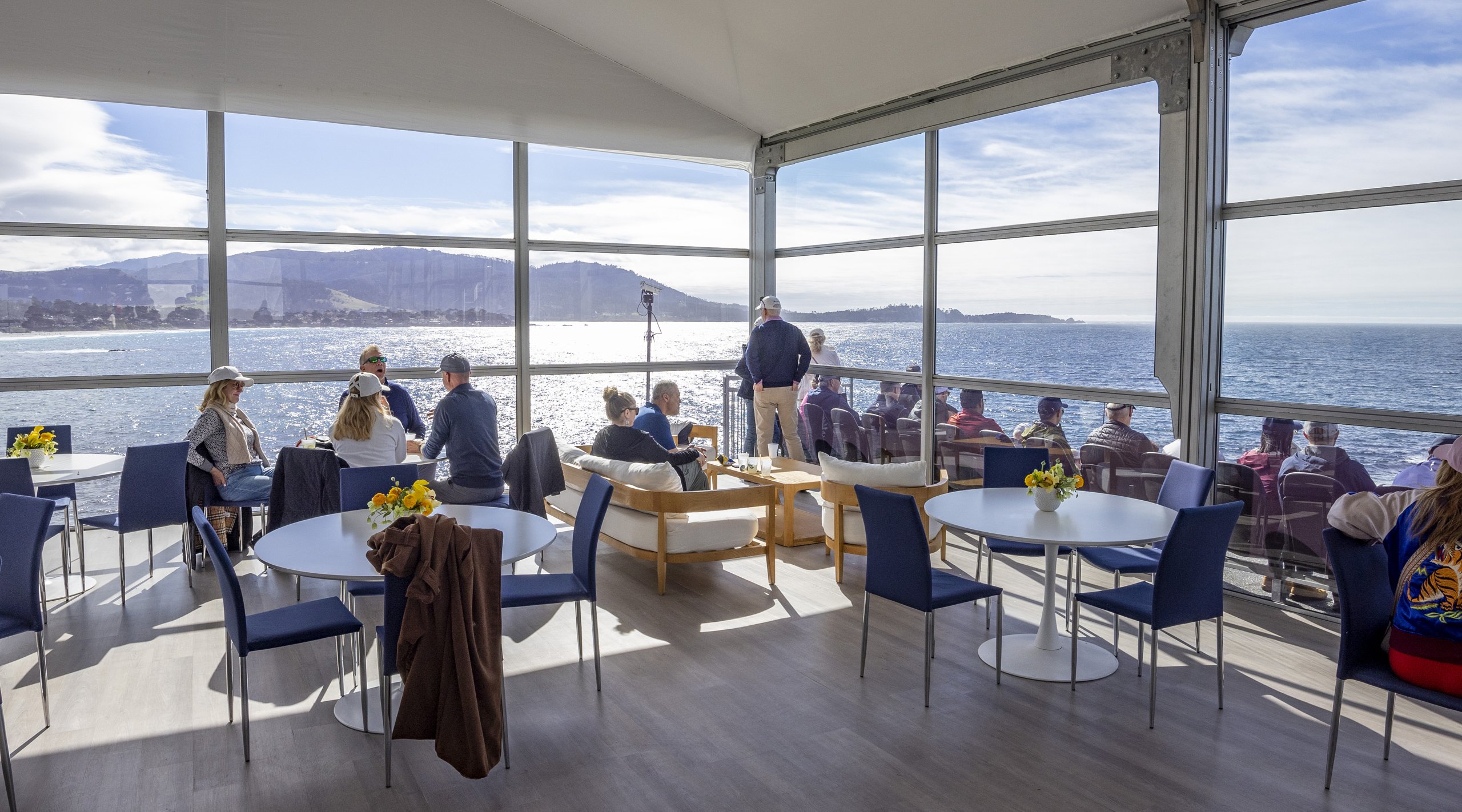 The interior of a sunny, glass-enclosed hospitality tent overlooking the ocean and coastline, furnished with modern white tables and blue chairs, and a small lounge area.