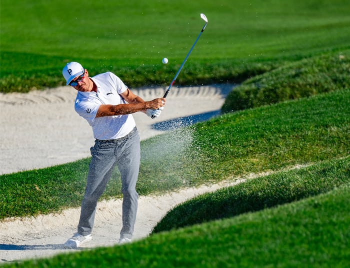 Golfer Rickie Fowler is mid-shot, hitting the ball out of a large sand bunker on a sunny day, kicking up a large spray of white sand and dust.